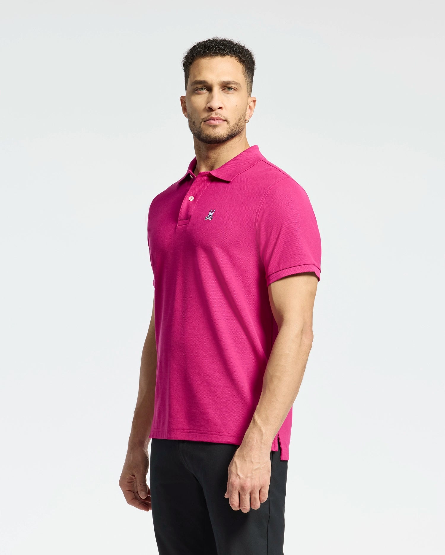 A man poses against a plain background in Psycho Bunny's bright pink MENS CLASSIC PIQUE POLO SHIRT - B6K001CRPC with an embroidered Bunny logo, paired with black pants. His short curly hair and neatly trimmed beard enhance his casual yet confident look.