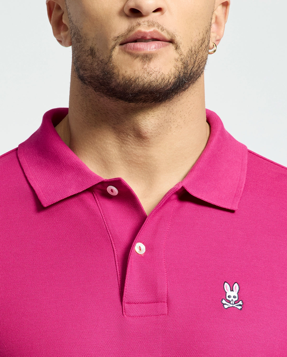 A person is wearing a Psycho Bunny Men's Classic Piqué Polo Shirt (B6K001CRPC) in bright pink, featuring an embroidered Bunny logo with crossbones on the chest. Made of soft Pima cotton, it includes a stylish collar and button details. The background is plain light gray.
