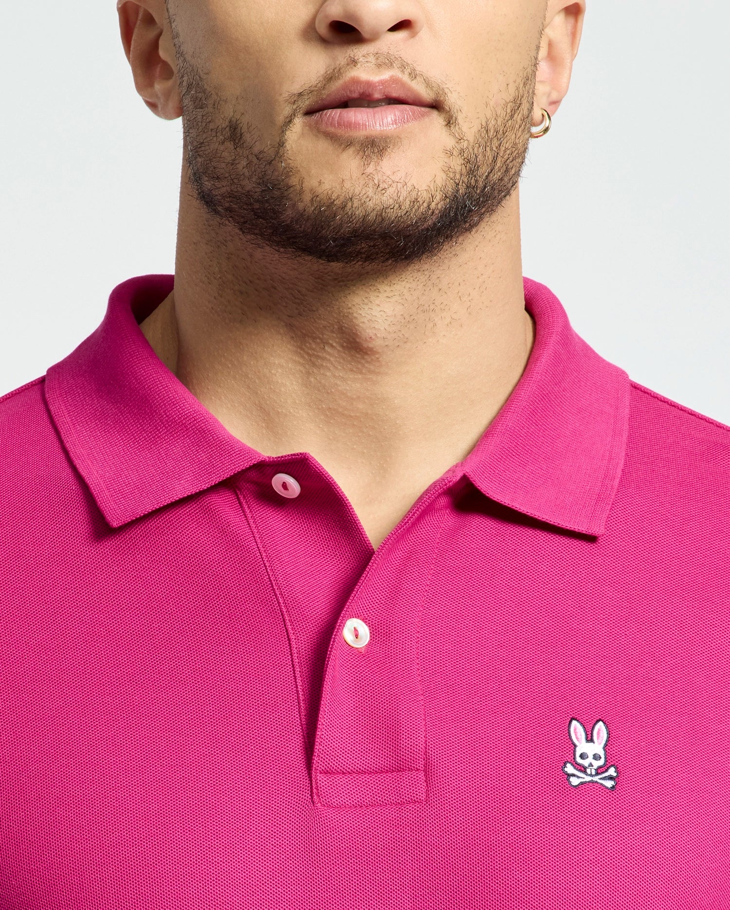 A person is wearing a Psycho Bunny Men's Classic Piqué Polo Shirt (B6K001CRPC) in bright pink, featuring an embroidered Bunny logo with crossbones on the chest. Made of soft Pima cotton, it includes a stylish collar and button details. The background is plain light gray.