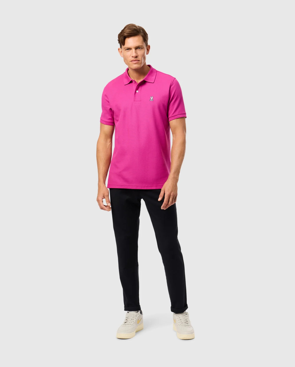 Dressed in Psycho Bunny's MENS CLASSIC PIQUE POLO SHIRT, made from 100% Pima cotton in Peru, featuring a small logo, the person pairs it with black pants and light sneakers. They pose against a plain background, looking at the camera.