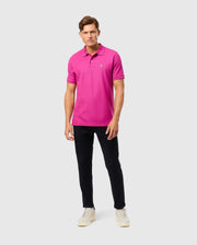 Dressed in Psycho Bunny's MENS CLASSIC PIQUE POLO SHIRT, made from 100% Pima cotton in Peru, featuring a small logo, the person pairs it with black pants and light sneakers. They pose against a plain background, looking at the camera.