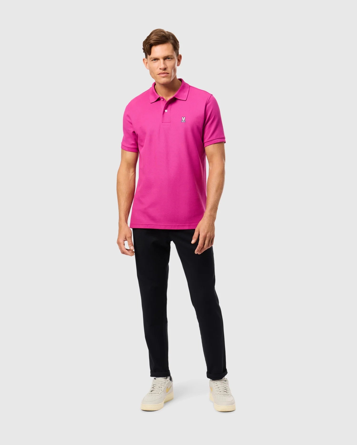 Dressed in Psycho Bunny's MENS CLASSIC PIQUE POLO SHIRT, made from 100% Pima cotton in Peru, featuring a small logo, the person pairs it with black pants and light sneakers. They pose against a plain background, looking at the camera.