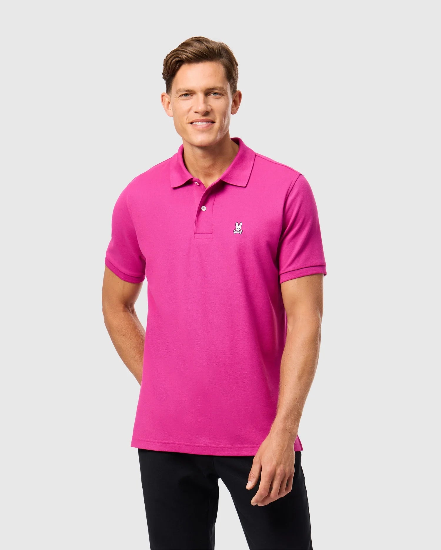 A person in a Psycho Bunny MENS CLASSIC PIQUE POLO SHIRT - B6K001D200, bright pink, made in Peru from 100% Pima cotton, stands against a plain gray background with short brown hair and smiles at the camera.