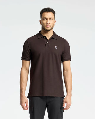 A man wearing a Psycho Bunny Mens Classic Pique Polo Shirt (B6K001D200) in dark brown, made from Pima cotton with mother-of-pearl buttons, stands against a light gray background. His short, curly hair enhances his direct gaze.