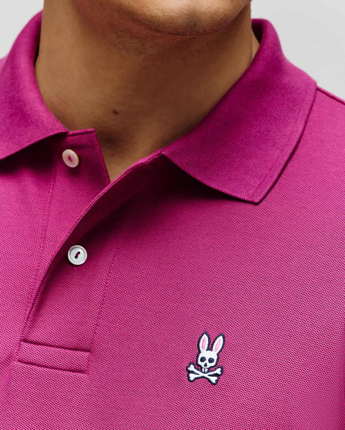 A close-up of a person wearing the Psycho Bunny MENS CLASSIC PIQUE POLO (B6K001F200) in magenta, made from soft Pima cotton, with white buttons and the embroidered Bunny logo with crossbones on the chest.