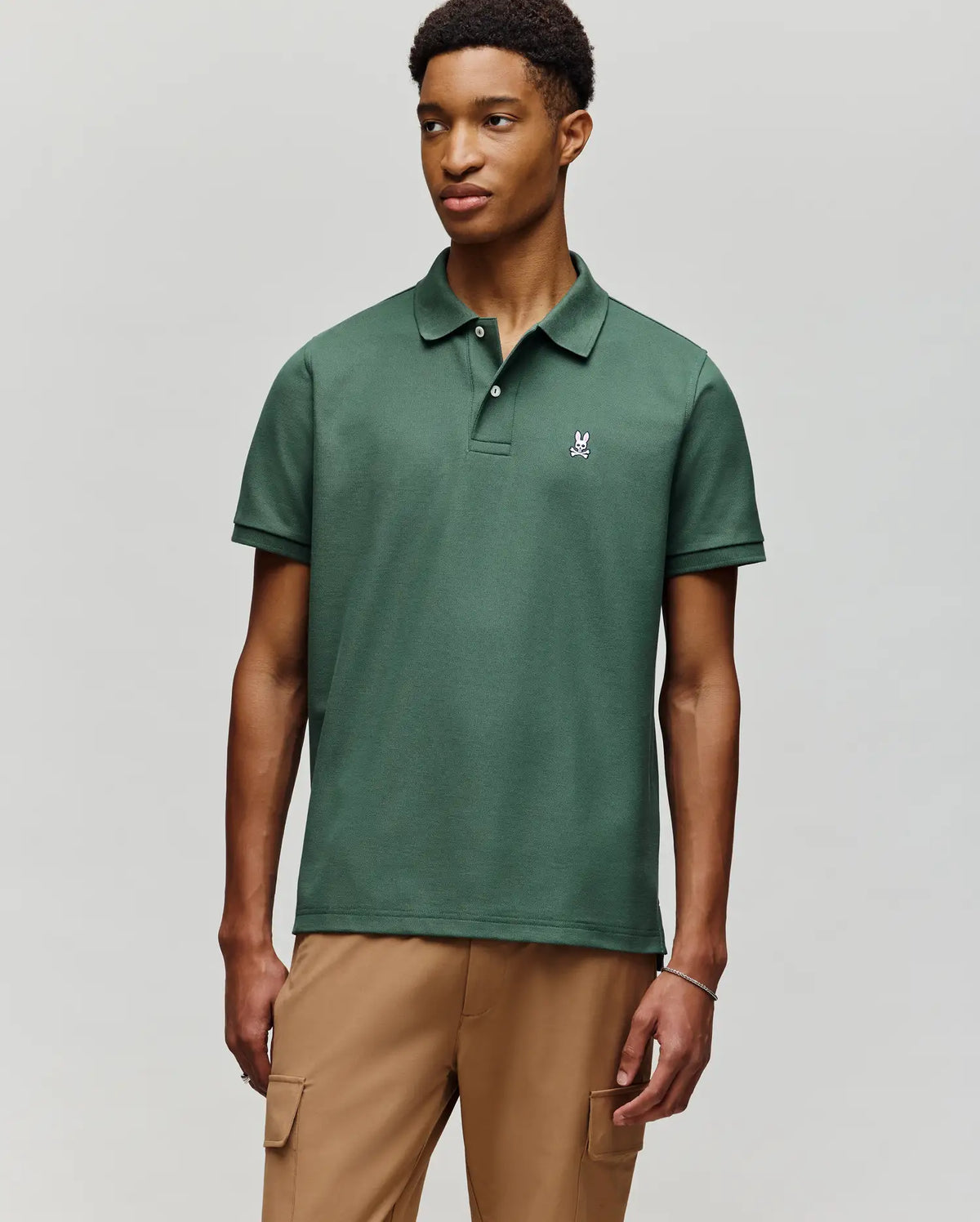 A young man wears the Psycho Bunny MENS CLASSIC PIQUE POLO - B6K001F200, featuring soft Pima cotton and an embroidered Bunny logo, paired with light brown cargo pants against a plain light gray background.
