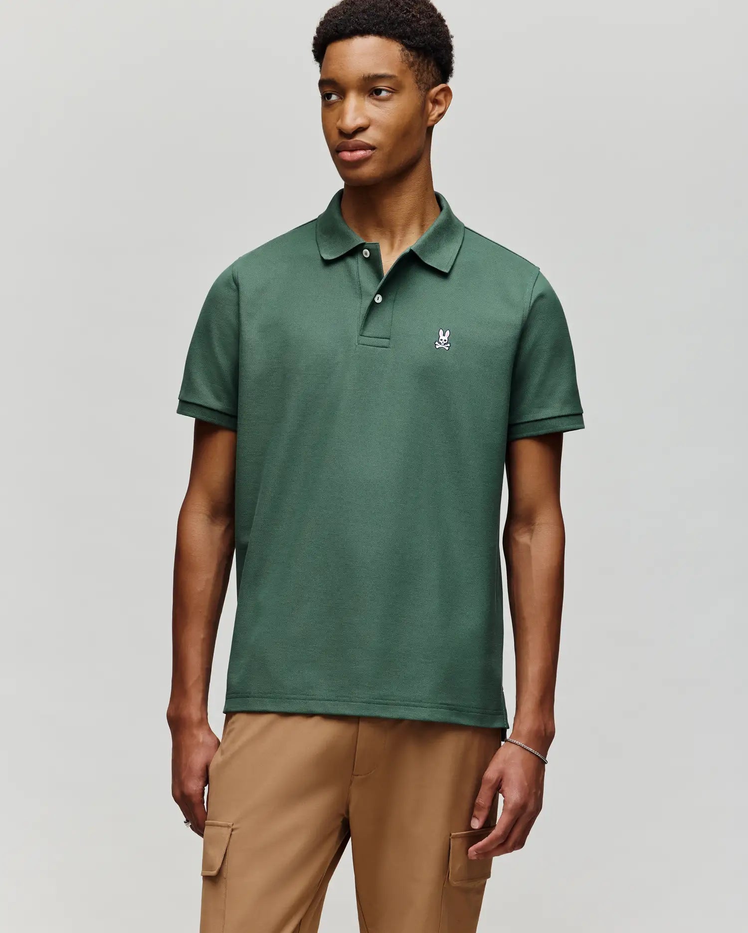 A young man wears the Psycho Bunny MENS CLASSIC PIQUE POLO - B6K001F200, featuring soft Pima cotton and an embroidered Bunny logo, paired with light brown cargo pants against a plain light gray background.