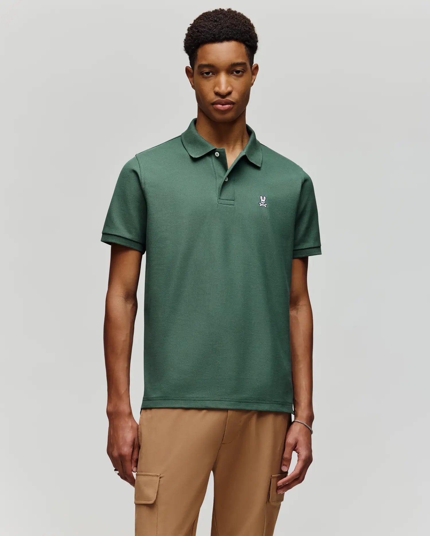 A man wears the Psycho Bunny MENS CLASSIC PIQUE POLO - B6K001F200, featuring an embroidered Bunny logo, with tan cargo pants against a light gray background.