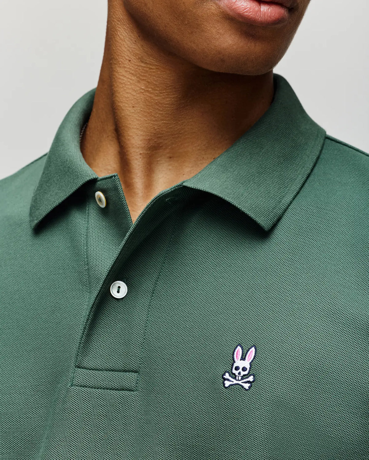 A person wears the Psycho Bunny MENS CLASSIC PIQUE POLO - B6K001F200 in green Pima cotton, with an embroidered pink-eared bunny above white crossbones logo on the left chest. Only the lower face and upper torso are visible.
