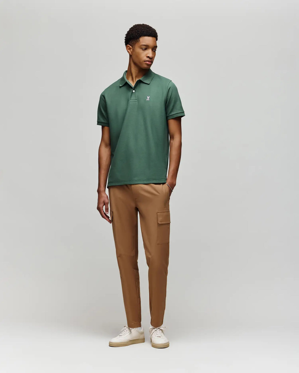 A man stands against a light gray background wearing a green Psycho Bunny MENS CLASSIC PIQUE POLO (B6K001F200) with an embroidered Bunny logo, tan cargo pants, and white sneakers. He looks slightly to the side in a relaxed pose.