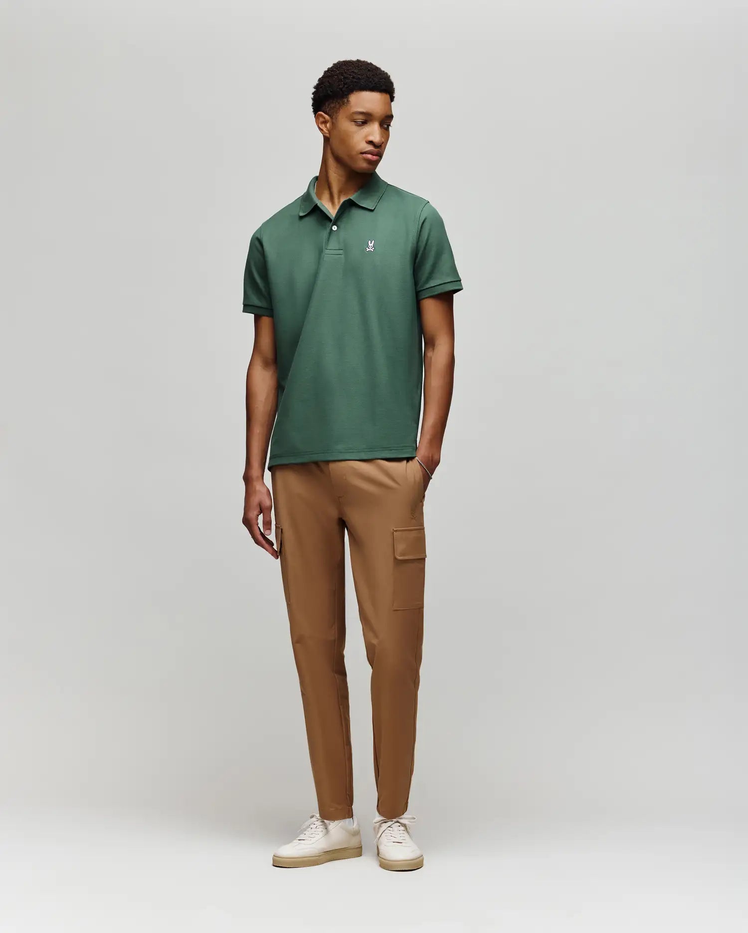 A man stands against a light gray background wearing a green Psycho Bunny MENS CLASSIC PIQUE POLO (B6K001F200) with an embroidered Bunny logo, tan cargo pants, and white sneakers. He looks slightly to the side in a relaxed pose.