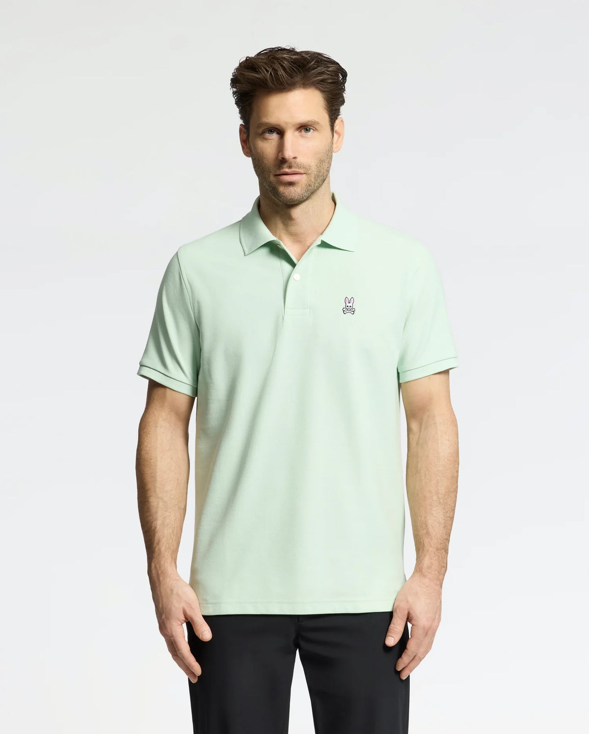 A man wearing the Psycho Bunny MENS CLASSIC PIQUE POLO (B6K001F200), made with soft Pima cotton and a small bunny logo on the chest, stands against a white background. He has short brown hair, black pants, and looks directly at the camera.