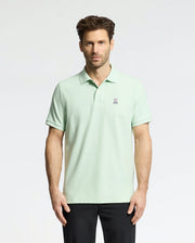 A man wearing the Psycho Bunny MENS CLASSIC PIQUE POLO (B6K001F200), made with soft Pima cotton and a small bunny logo on the chest, stands against a white background. He has short brown hair, black pants, and looks directly at the camera.