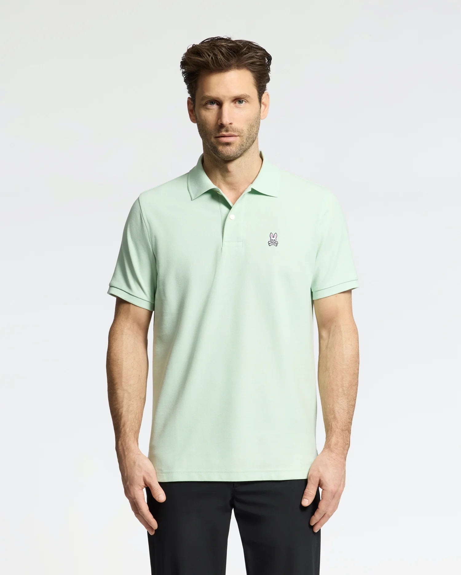 A man wearing the Psycho Bunny MENS CLASSIC PIQUE POLO (B6K001F200), made with soft Pima cotton and a small bunny logo on the chest, stands against a white background. He has short brown hair, black pants, and looks directly at the camera.