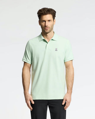 A man wearing the Psycho Bunny MENS CLASSIC PIQUE POLO (B6K001F200), made with soft Pima cotton and a small bunny logo on the chest, stands against a white background. He has short brown hair, black pants, and looks directly at the camera.