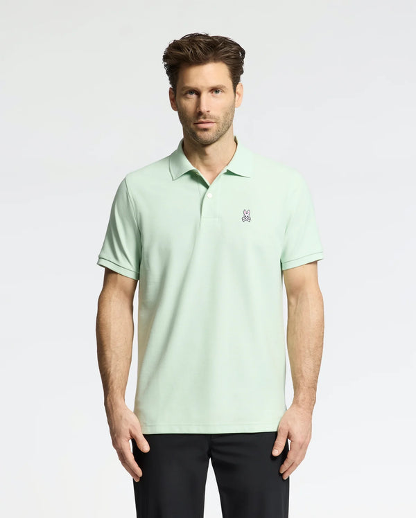 A man wearing the Psycho Bunny MENS CLASSIC PIQUE POLO (B6K001F200), made with soft Pima cotton and a small bunny logo on the chest, stands against a white background. He has short brown hair, black pants, and looks directly at the camera.