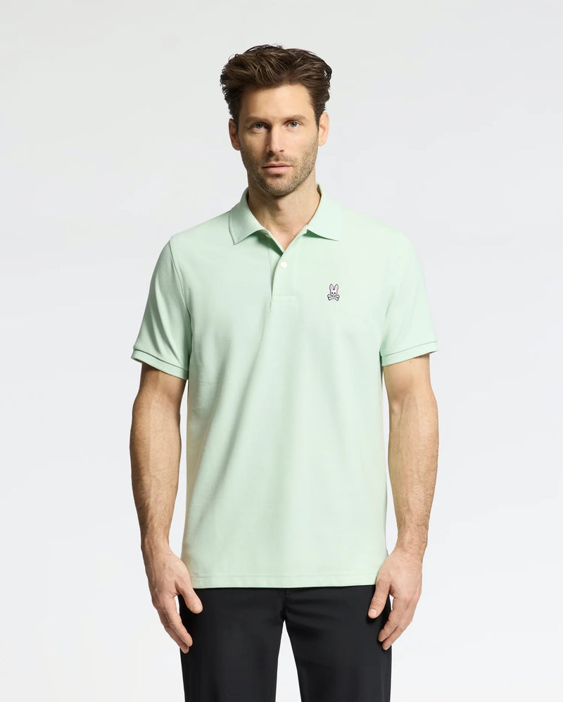 A man wearing the Psycho Bunny MENS CLASSIC PIQUE POLO (B6K001F200), made with soft Pima cotton and a small bunny logo on the chest, stands against a white background. He has short brown hair, black pants, and looks directly at the camera.