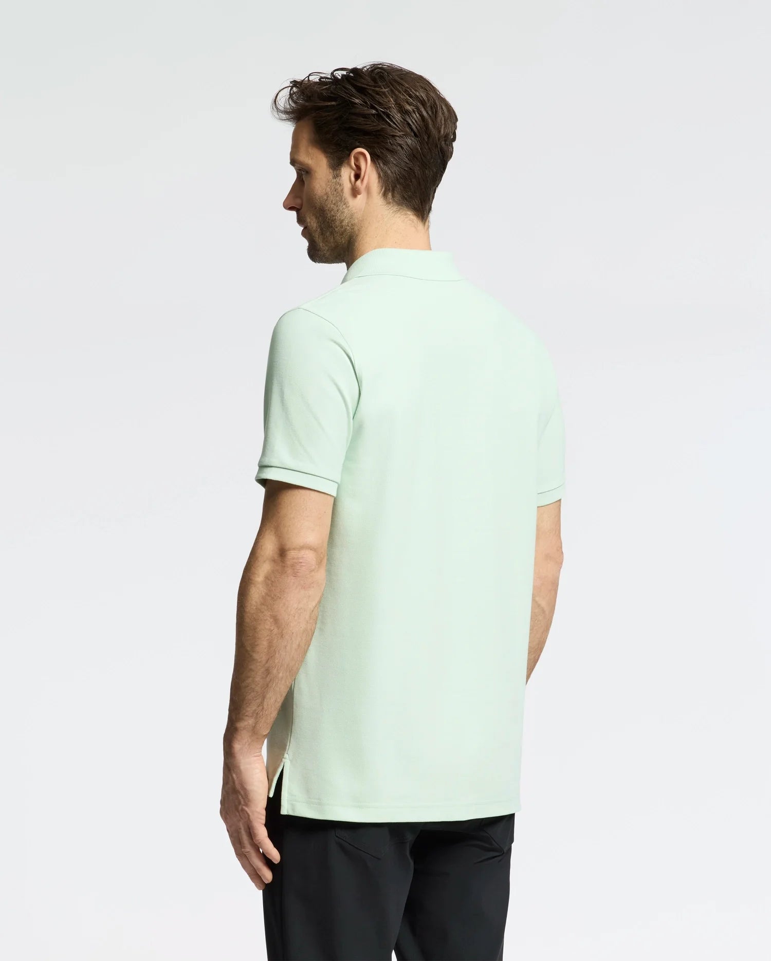 A man with short brown hair models the Psycho Bunny Men's Classic Piqué Polo (B6K001F200) in light green. Made from soft Pima cotton, it features a subtle bunny logo. He pairs it with black pants against a plain light gray background, facing away from the camera.