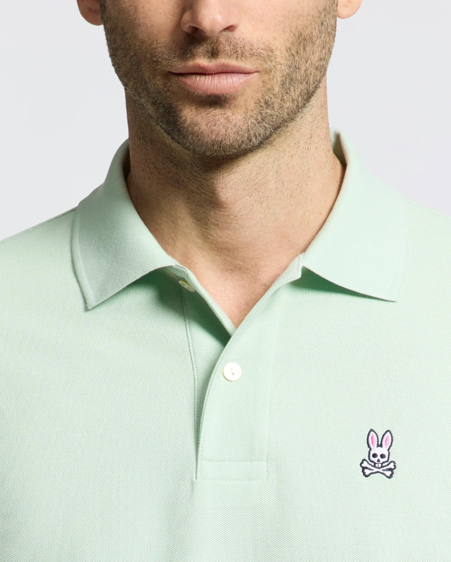 A man wears a Psycho Bunny MENS CLASSIC PIQUE POLO (B6K001F200) in light green, highlighted by its pink bunny and crossbones logo. Made from Pima cotton, it features a comfortable collar with two buttons undone against a plain white backdrop.