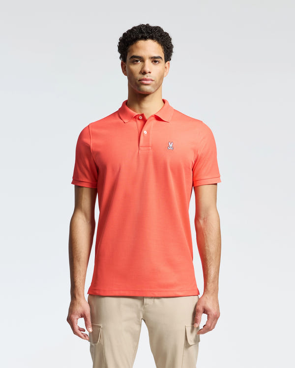 A man stands against a neutral background, wearing beige pants and a Psycho Bunny MENS CLASSIC PIQUE POLO - B6K001F200, made from soft Pima cotton with a small bunny logo on the chest, looking directly at the camera.