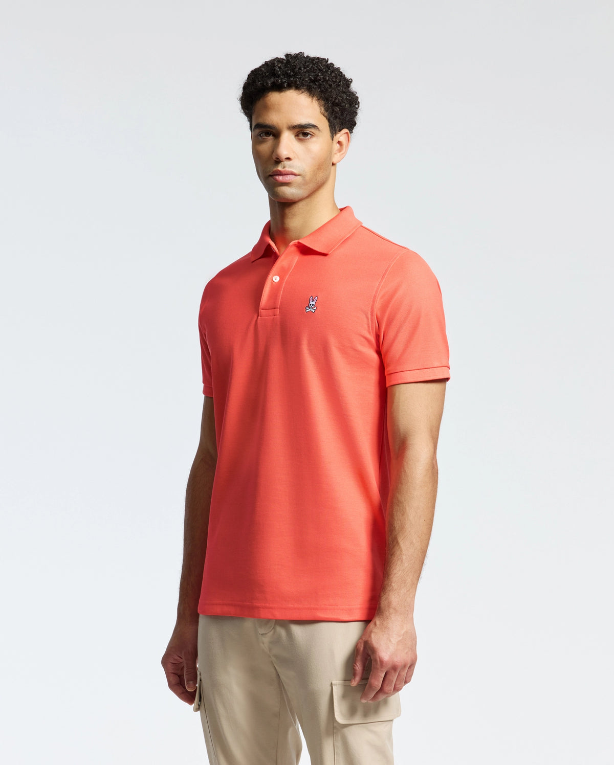 A person stands wearing the Psycho Bunny MENS CLASSIC PIQUE POLO - B6K001F200 in bright orange with an embroidered bunny logo on the chest, paired with light beige pants against a plain, light-colored background.