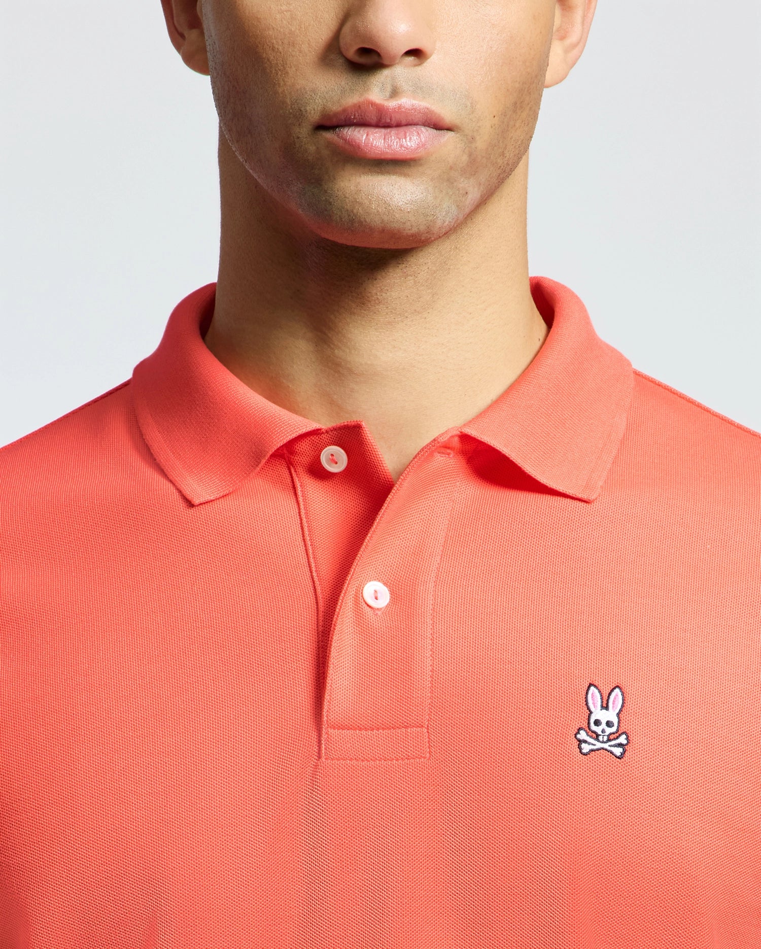 A man models the Psycho Bunny MENS CLASSIC PIQUE POLO - B6K001F200 in coral, made from soft Pima cotton. The upper torso shot showcases the small embroidered bunny with crossbones on the left chest.