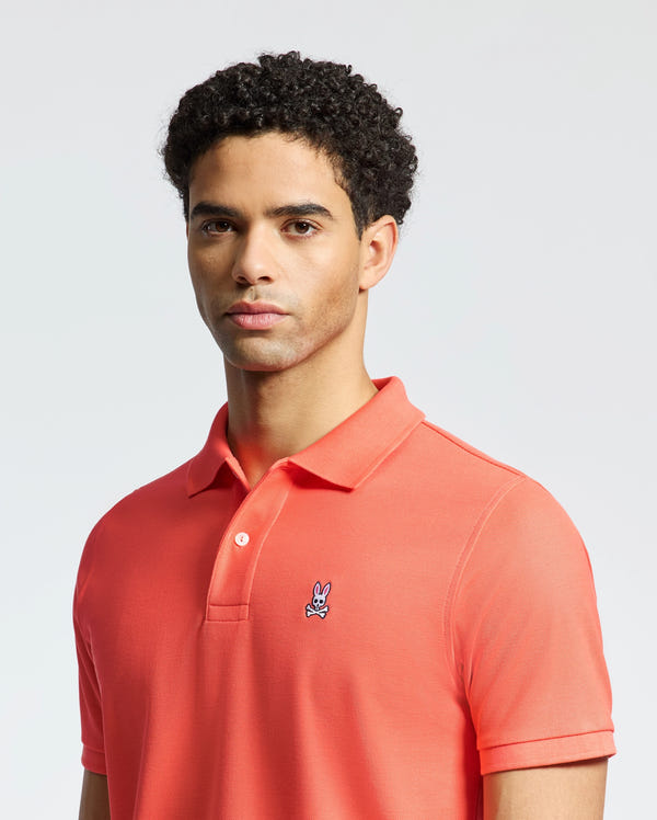 A person with curly hair wears a Psycho Bunny MENS CLASSIC PIQUE POLO - B6K001F200 in coral, crafted from soft Pima cotton with an embroidered bunny logo, set against a plain neutral background.