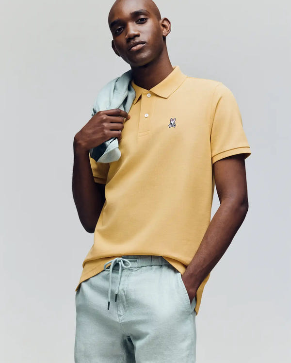 A man in a Psycho Bunny MENS CLASSIC PIQUE POLO (B6K001F200), yellow with an embroidered Bunny logo, and light gray drawstring pants poses with a sweater over his shoulder against a plain background.