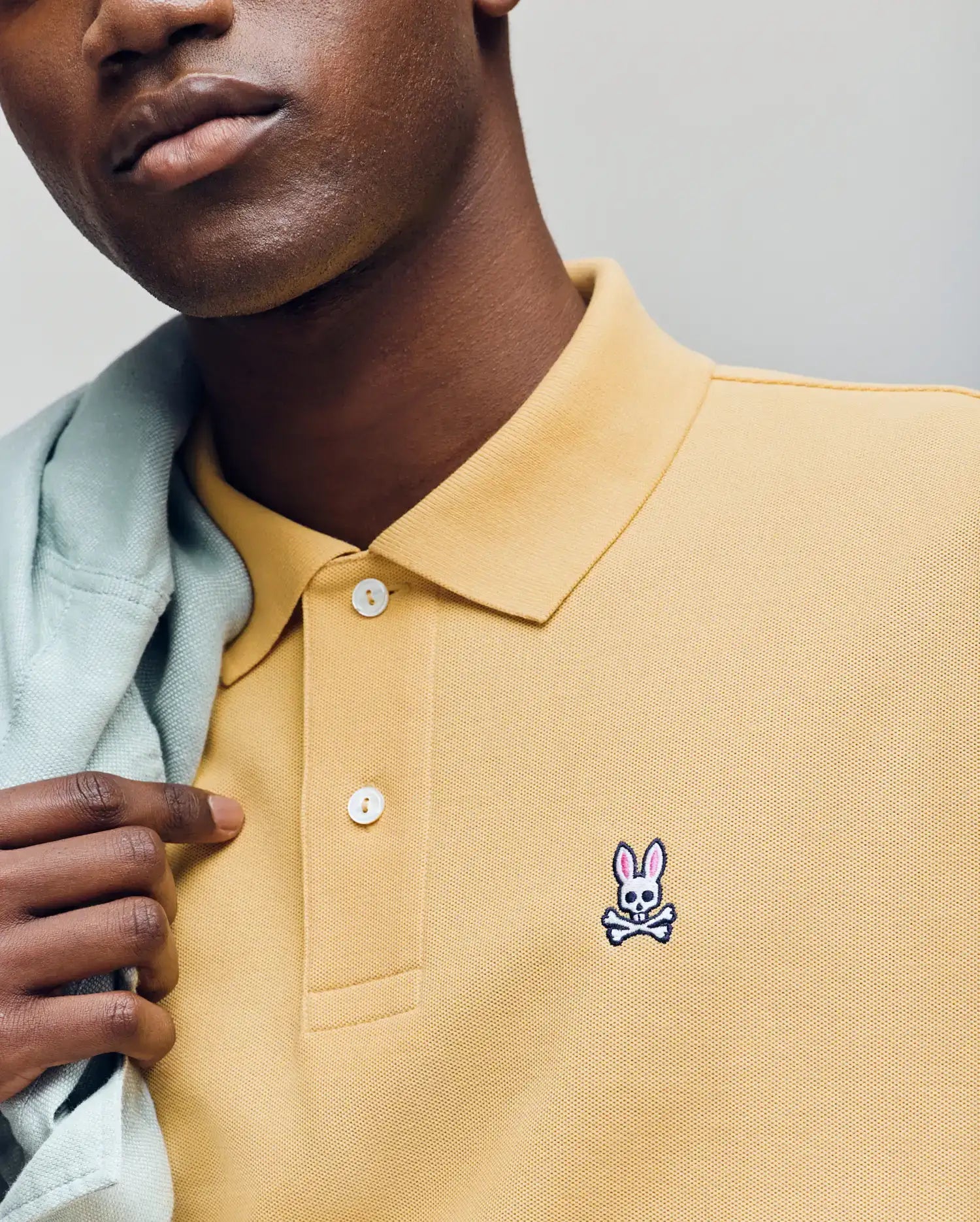Wearing Psycho Bunny's MENS CLASSIC PIQUE POLO - B6K001F200 in yellow, made from Pima cotton and featuring the embroidered Bunny logo, a person holds a light jacket over their shoulder with only the lower half of their face visible.