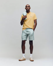 A man stands against a plain background wearing a yellow Psycho Bunny MENS CLASSIC PIQUE POLO - B6K001F200 with an embroidered Bunny logo, light green shorts and matching jacket, plus beige sneakers with white socks.