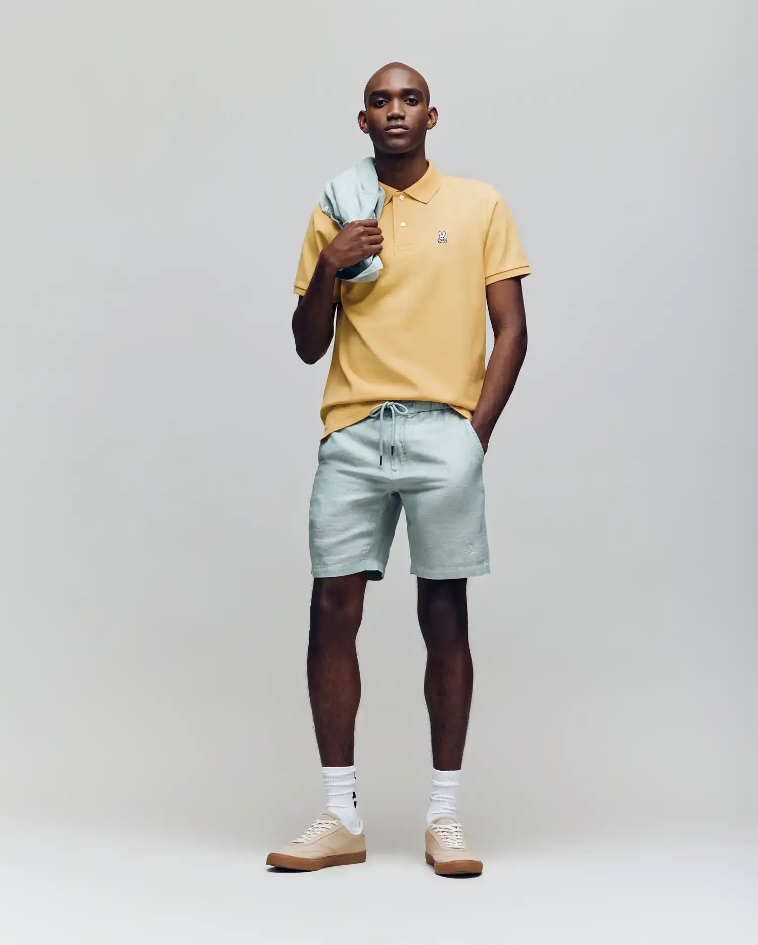 A man stands against a plain background wearing a yellow Psycho Bunny MENS CLASSIC PIQUE POLO - B6K001F200 with an embroidered Bunny logo, light green shorts and matching jacket, plus beige sneakers with white socks.