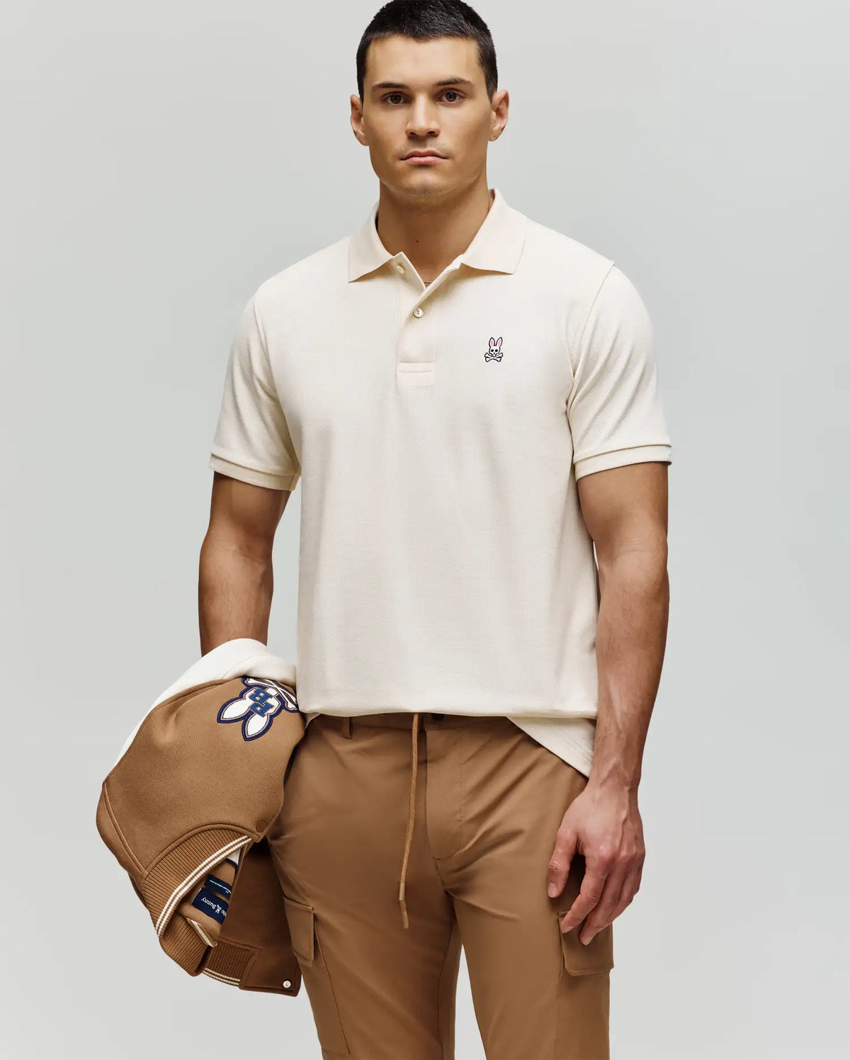 A man with short dark hair wears a Psycho Bunny MENS CLASSIC PIQUE POLO SHIRT (B6K001F200) in cream and tan pants, holding a tan jacket with blue and white design over his left arm, standing against a plain light background.