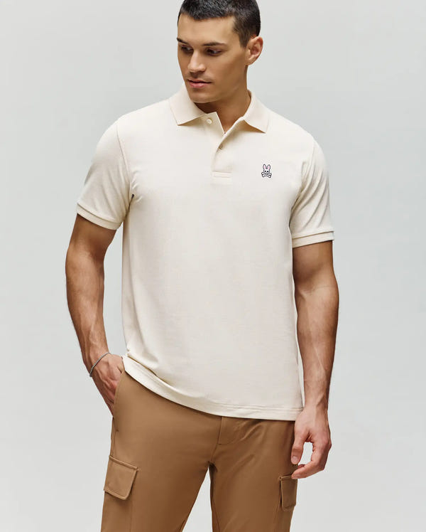 A man in a Psycho Bunny MENS CLASSIC PIQUE POLO SHIRT - B6K001F200 with an embroidered Bunny logo and tan cargo pants stands with one hand in his pocket, looking down and to the side against a plain background.
