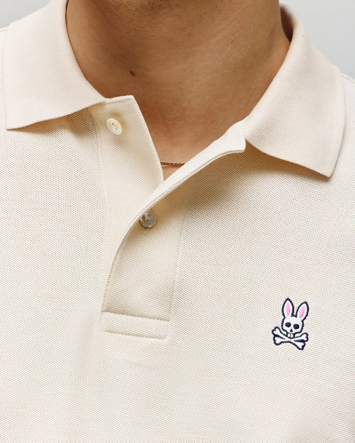 Close-up of a person wearing the Psycho Bunny MENS CLASSIC PIQUE POLO SHIRT - B6K001F200 in cream, featuring two buttons undone and an embroidered Bunny logo with pink ears and crossbones on the left chest.