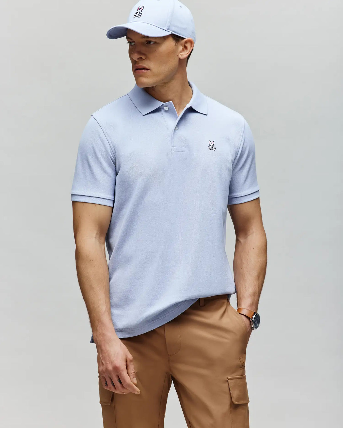 A man in a Psycho Bunny MENS CLASSIC PIQUE POLO - B6K001F200 in light blue with a small bunny logo, paired with a matching cap and tan cargo pants, stands against a gray background looking to the side.