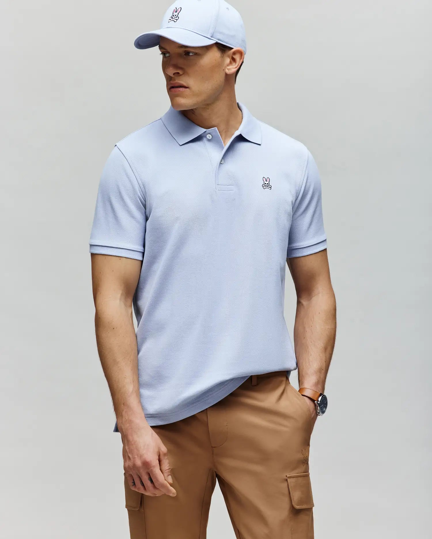 A man in a Psycho Bunny MENS CLASSIC PIQUE POLO - B6K001F200 in light blue with a small bunny logo, paired with a matching cap and tan cargo pants, stands against a gray background looking to the side.