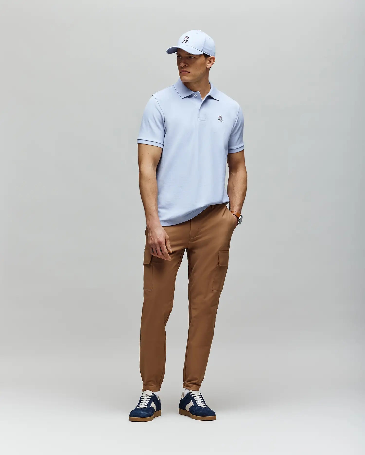 A man stands against a light gray background wearing a light blue Psycho Bunny MENS CLASSIC PIQUE POLO - B6K001F200, tan cargo pants, navy and white sneakers, and a matching light blue baseball cap.