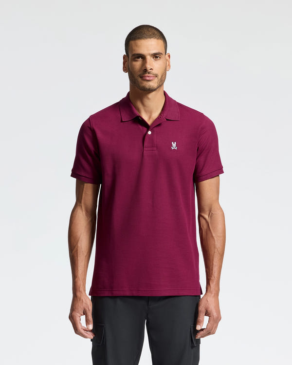 A man in a maroon Men's Classic Piqué Polo Shirt - B6K001Z1PC by Psycho Bunny, with an embroidered Bunny logo on the chest, stands against a white background. He looks directly at the camera with a neutral expression, his hands by his sides.