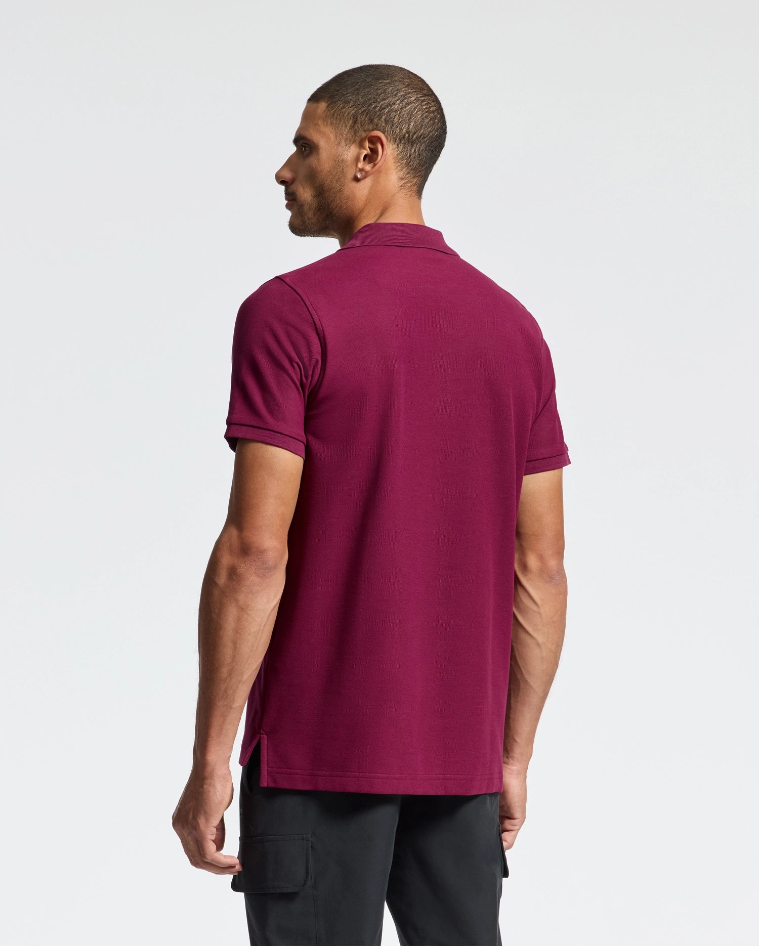 A man with short dark hair stands facing away. He's wearing a Psycho Bunny maroon MENS CLASSIC PIQUE POLO SHIRT, B6K001Z1PC, made of soft Pima cotton with an embroidered Bunny logo, paired with black pants against a plain white background.