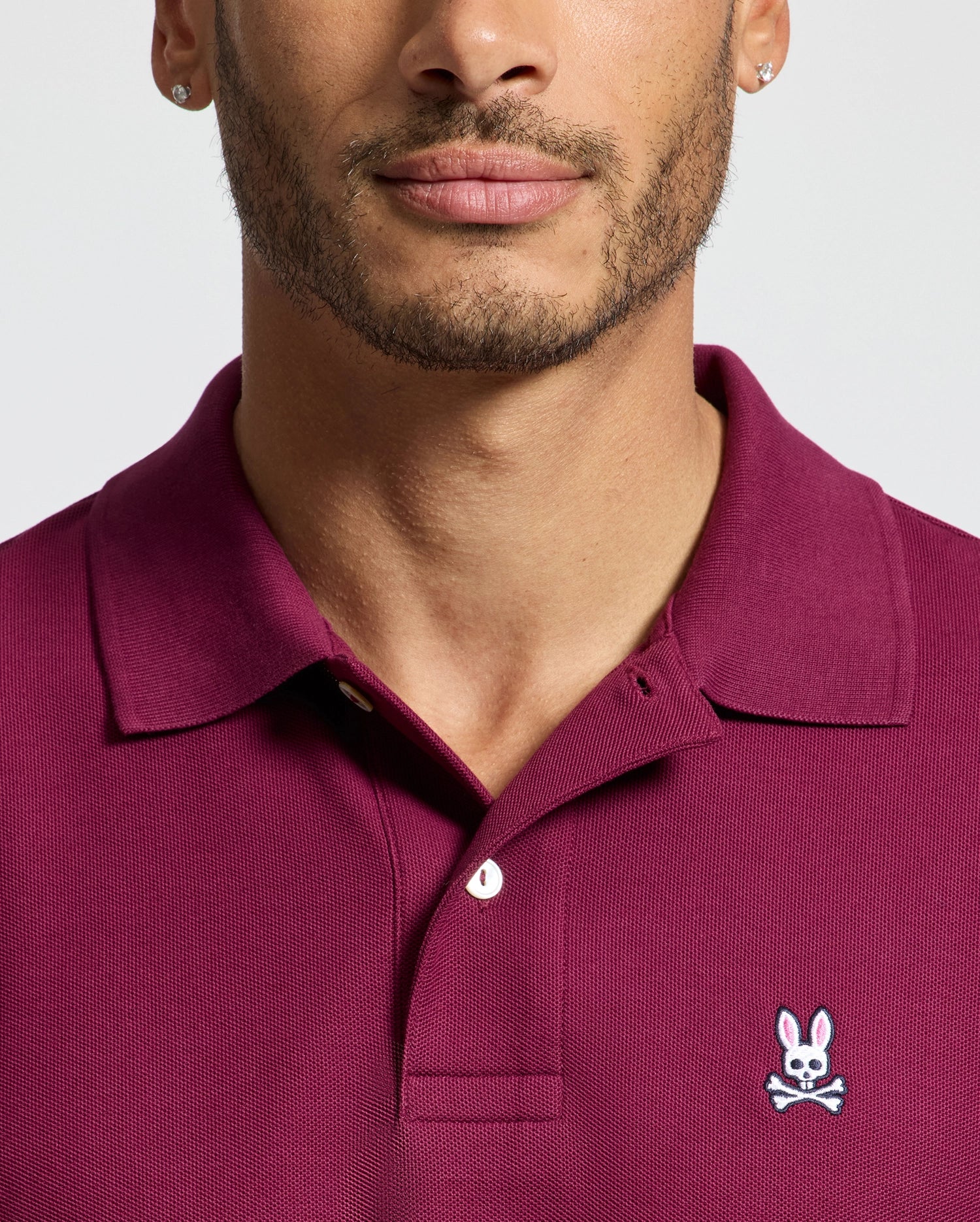 A person wears a Psycho Bunny MENS CLASSIC PIQUE POLO SHIRT (B6K001Z1PC) in maroon, featuring an embroidered Bunny logo with a cartoon skull and bunny ears on the chest. Crafted from luxurious Pima cotton, the focus is on the upper body and shirt against a plain light gray background.