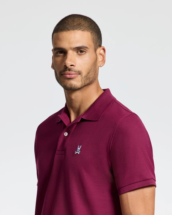 A person in a maroon Psycho Bunny Men's Classic Piqué Polo Shirt (B6K001Z1PC) with an embroidered Bunny logo stands against a light background, facing slightly to the side with short hair and a neutral expression.