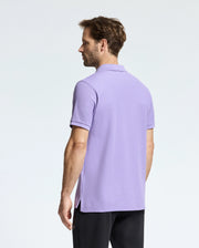 A man with short brown hair stands looking away, wearing the light purple Psycho Bunny MENS CLASSIC PIQUE POLO SHIRT - B6K001Z1PC with a subtle bunny logo, paired with black pants. He is set against a plain white background.