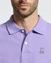 A man sports a Psycho Bunny Classic Pique Polo Shirt (B6K001Z1PC) in lavender, crafted from soft Pima cotton with a distinctive bunny and crossed bones logo on the chest, emphasizing its design and texture. His face is partially visible, showcasing his beard.