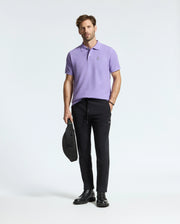 A man in a light purple Psycho Bunny Mens Classic Pique Polo Shirt - B6K001Z1PC with a subtle bunny logo and black pants stands holding a black bag, finishing his look with sleek black boots against a plain white backdrop.