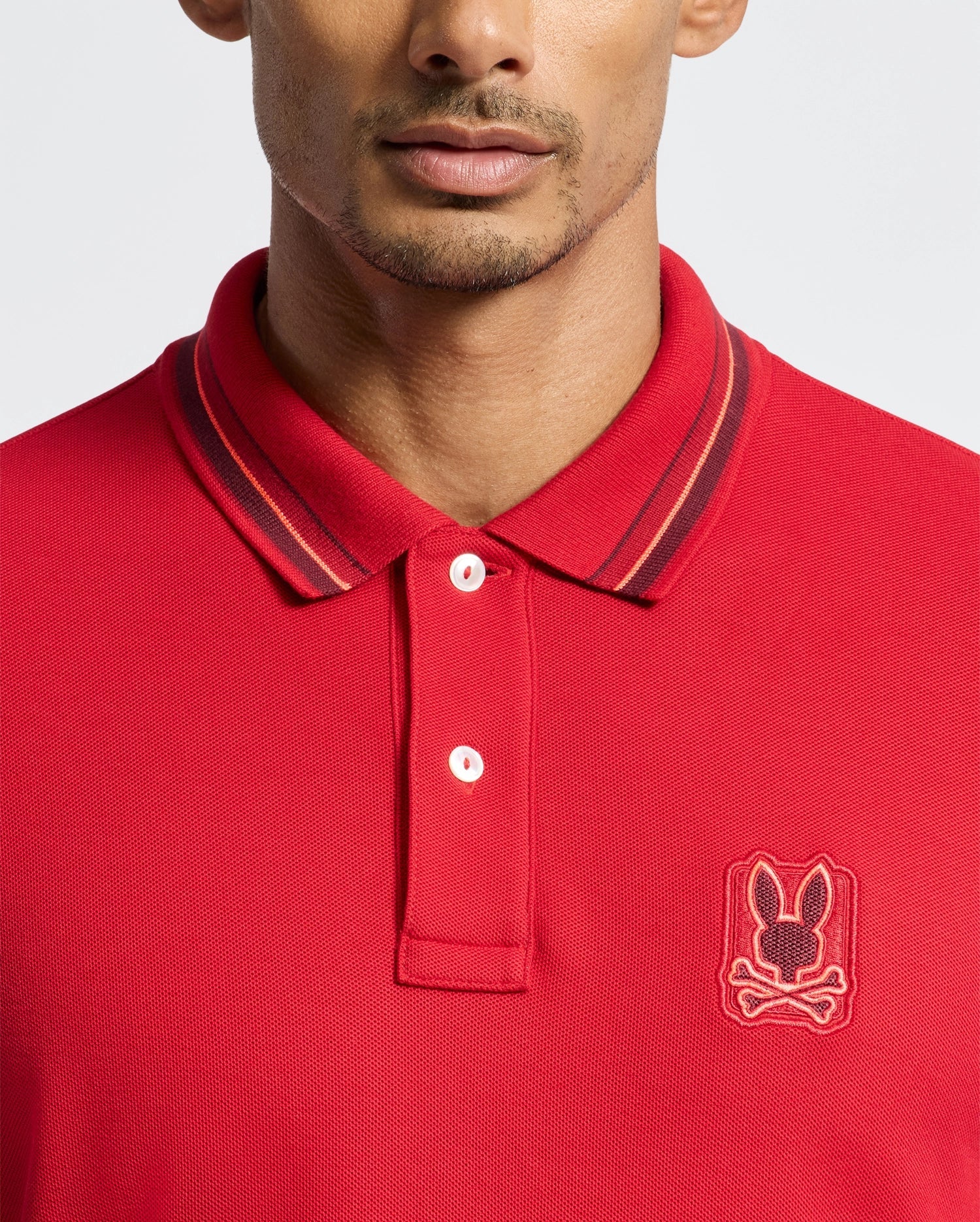 A person is dressed in the Psycho Bunny Men's Pierce Pique Polo (B6K201E200), which features a vibrant red color made from premium Pima cotton. The shirt is highlighted with dark striped detailing on the collar and adorned with an embroidered bunny skull logo on the chest. The polo also includes two fastened buttons, set against a simple, light-colored background.