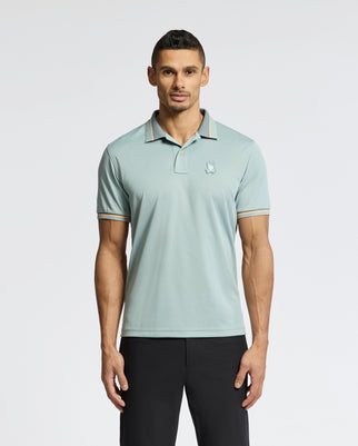 A man with short dark hair stands against a plain white background wearing the Psycho Bunny MENS STRATTON SPORT PIQUE POLO - B6K202E200 in light green with a subtle chest logo, contrasting collar and sleeve trim, and black pants. The polo features moisture-wicking fabric with a slight sheen.