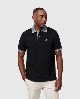 A man stands against a neutral background wearing the Psycho Bunny Men's Southport Pique Polo Shirt in black, which features a small embroidered logo and contrasting melange collar and sleeve trim. He pairs it with light pants, and his hands rest in his pockets.