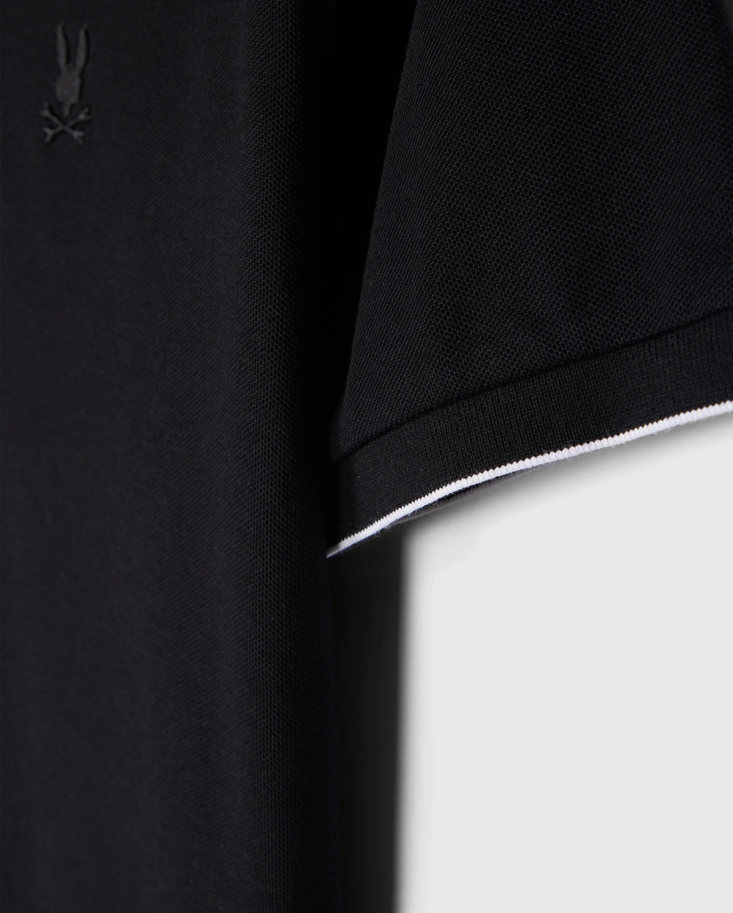 Close-up of a Psycho Bunny MENS EAST HILLS JOHNNY COLLAR POLO SHIRT - B6K331B200 sleeve with a subtle embroidered logo on the chest. The sleeve boasts a clean, minimalistic design featuring a single white stripe along the edge. Its solid pattern and neutral background enhance its timeless appeal.