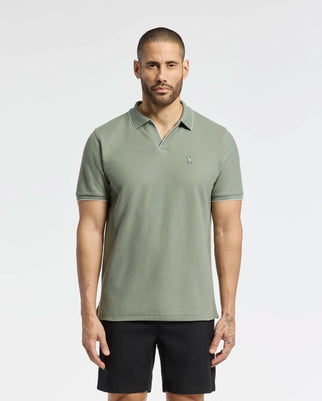 A bearded man with short hair and a right-hand tattoo stands against a white background, wearing Psycho Bunny's light green MENS EAST HILLS JOHNNY COLLAR POLO - B6K331B200, made of Pima cotton with an embroidered logo, paired with black shorts.