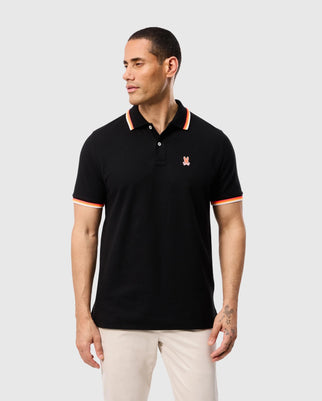 A person stands looking to the side, wearing a black Psycho Bunny Granbury Pique Polo (style code: B6K400C200) made from diamond-knit Pima cotton. The shirt showcases red and white striped detailing on the rib knit collar and sleeves, along with a small embroidered logo on the chest against a light gray background.